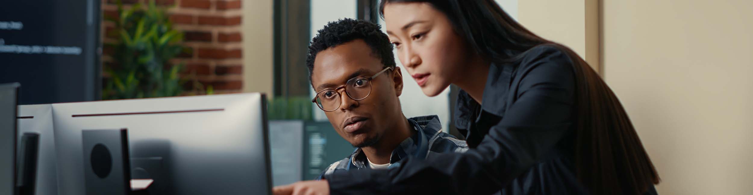 Two software developers coming at desk and sitting down holding laptop with coding interface and pointing at computer screen. Programmers team discussing algorithms looking at monitors with code.